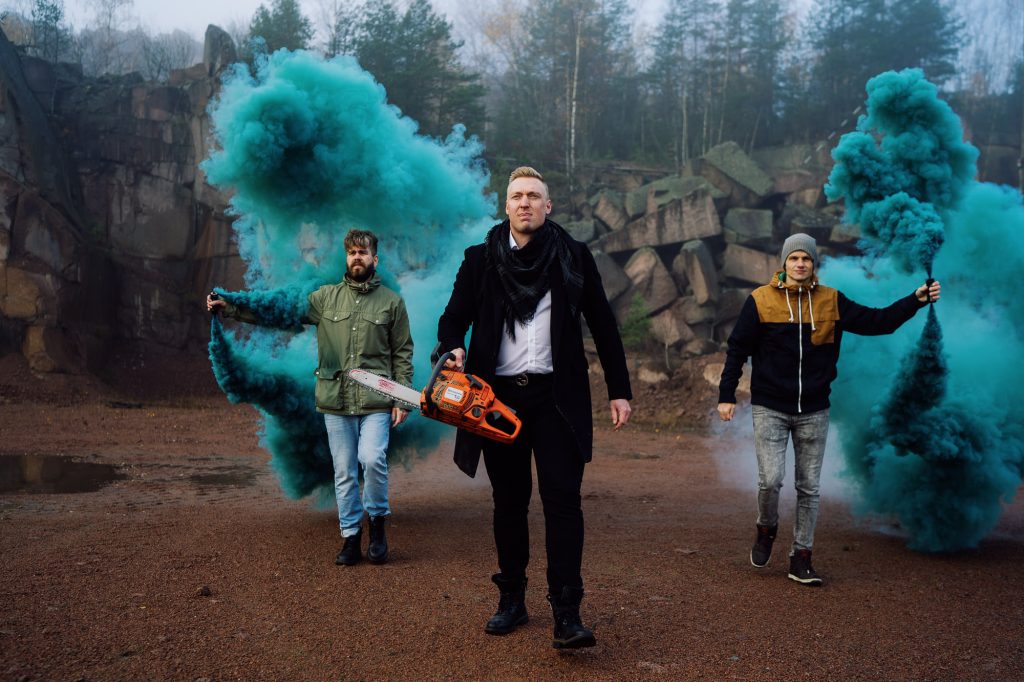Louhos Digitalin kolme osakasta louhoksella, takana ovat Petteri Poukka ja Juhani koskinen ja pitävät sinisiä savupatruunoita kädessään, keskellä Iiro Junnila ja moottorisaha.