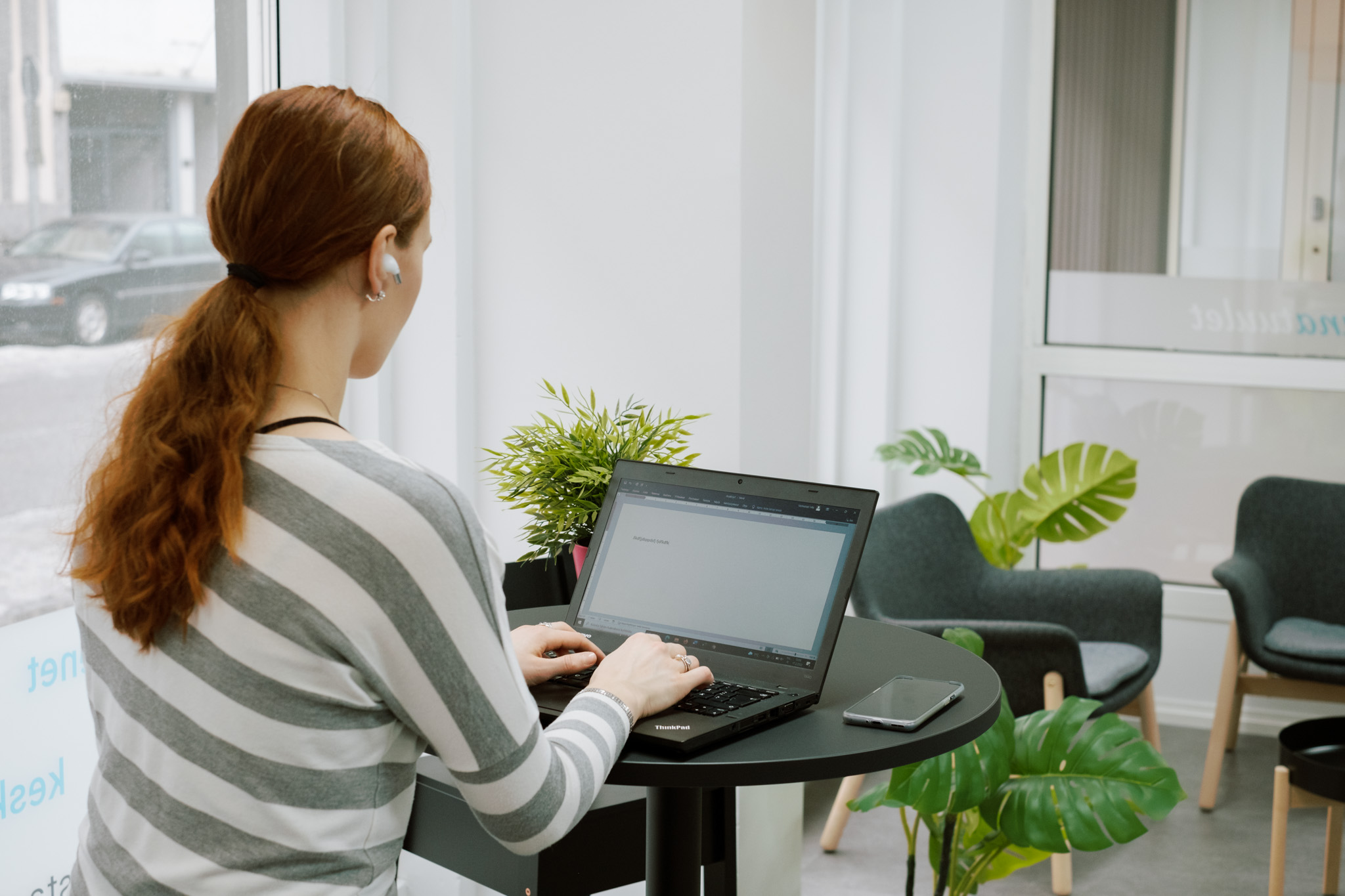 Nainen istuu pienen pyöreän pöydän ääressä ja kirjoittaa tietokoneella, hänellä on kuparinväriset pitkät hiukset ja kuva on otettu hänen selkänsä takaa.