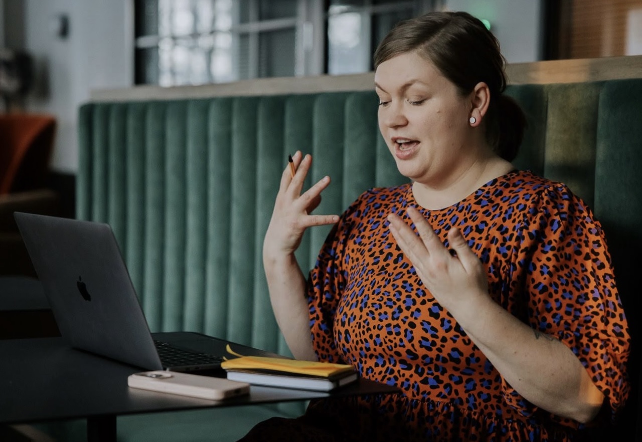 Tiina istuu tietokoneensa ääressä ja puhuu kädet edessään kämmenet kohti kasvoja.